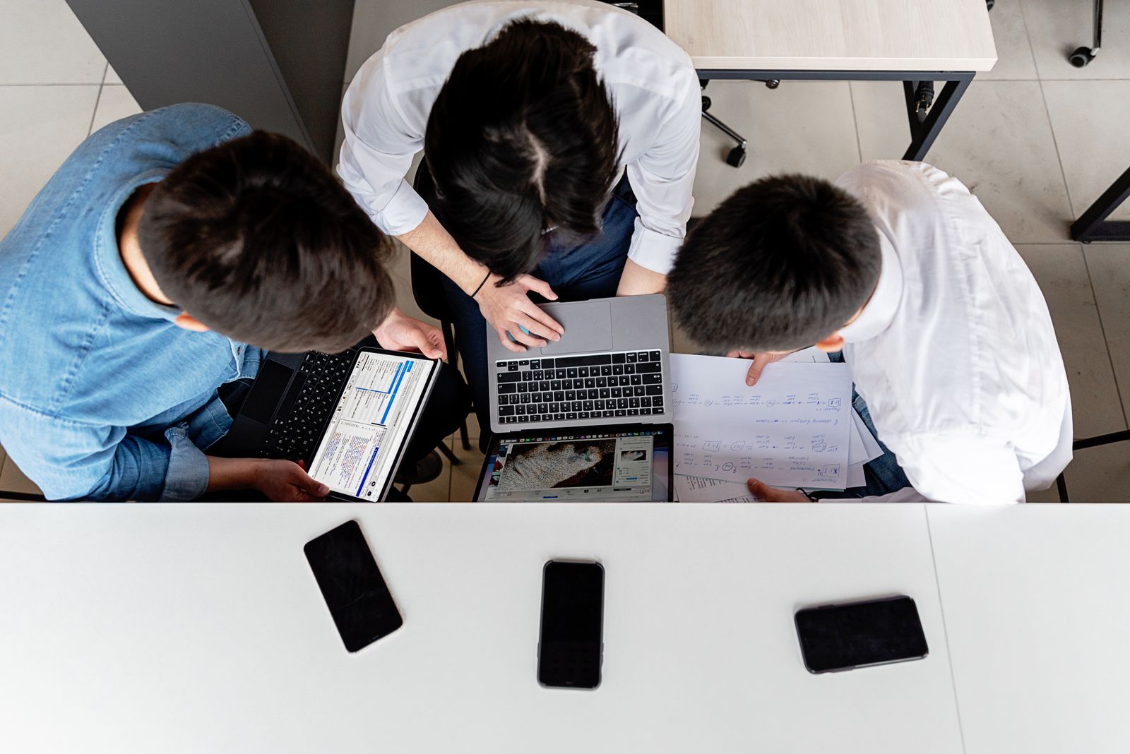 Three colleagues working together on laptops at an office desk with documents.