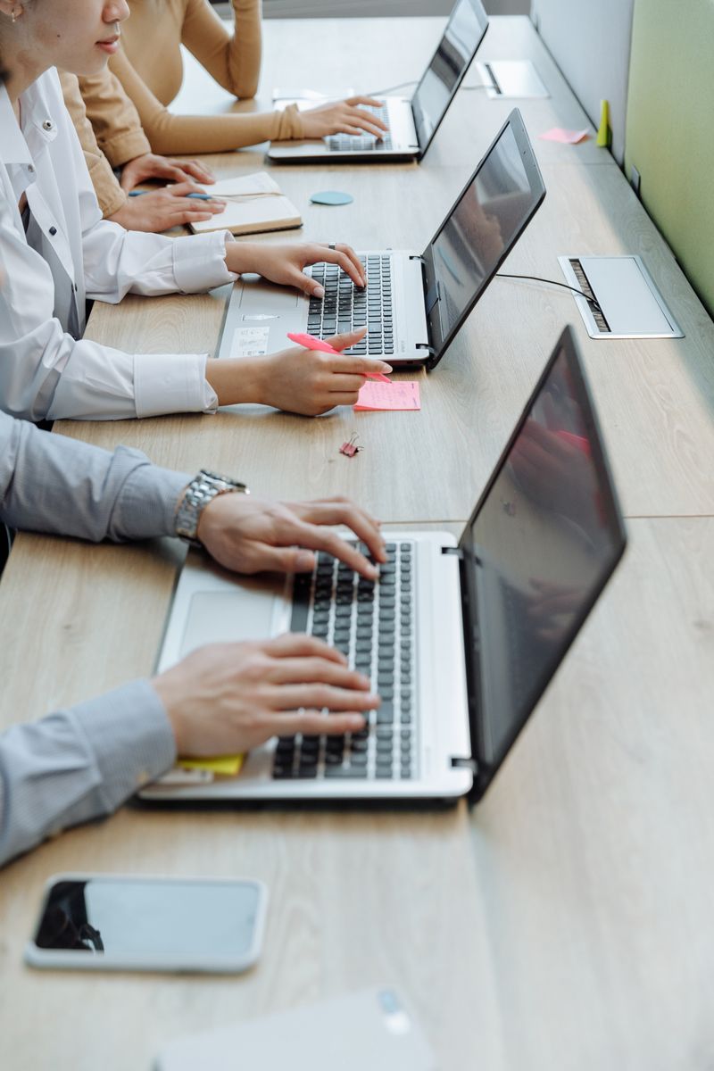 Group of office workers typing on laptops at a shared workspace desk.