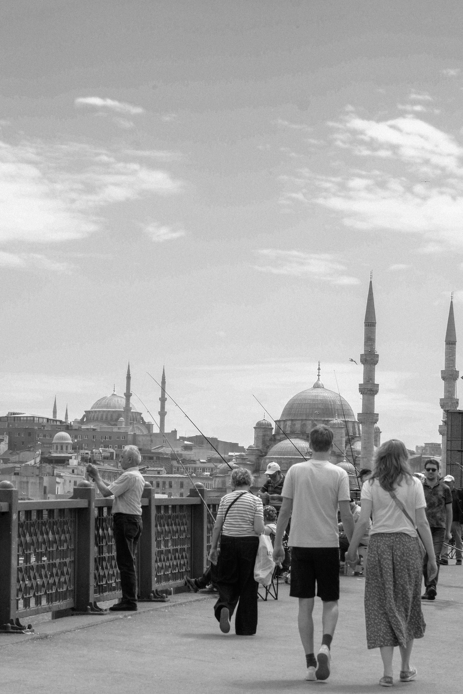 People walking on Galata Bridge in Istanbul, with domes and minarets in the background.