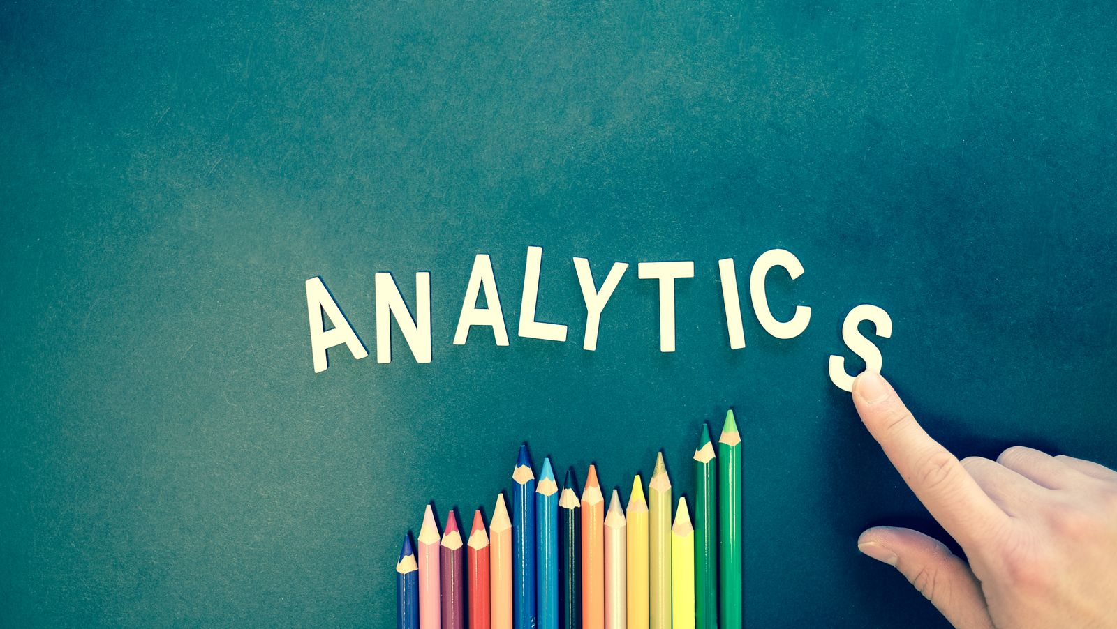Colored pencils and letters spelling &lsquo;ANALYTICS&rsquo; on a textured background with a hand.